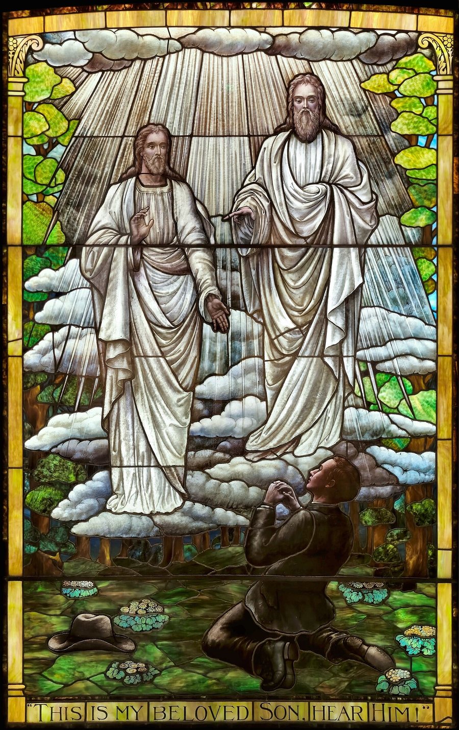 Stained glass: the Father and the Son standing in light above Joseph Smith, who kneels in the grove with his hat on the ground; the banner reads This is my beloved Son. Hear him!